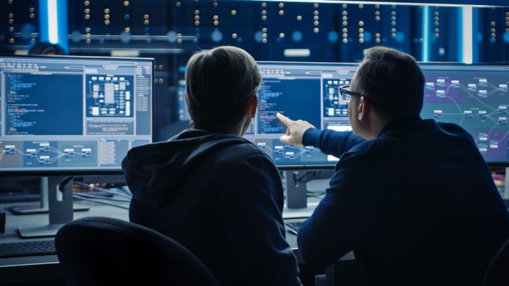 Two businessmen study their computer screens to check for security threats. Artificial intelligence and cyber crime can be used by hackers to gain powerful leverage over businesses.